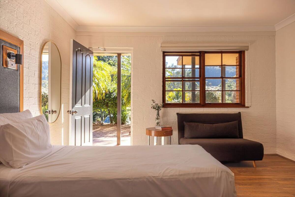 A bedroom with a bed, a bedside table with a book and a vase, a window with a view of greenery, and a door leading outside.