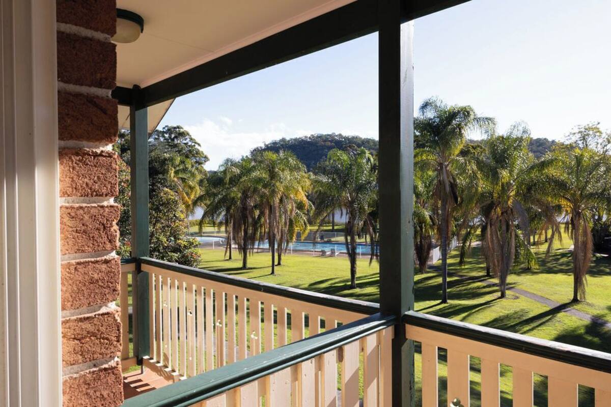 A balcony with a view of a well-maintained lawn and palm trees, a brick wall on one side, and a clear sky.