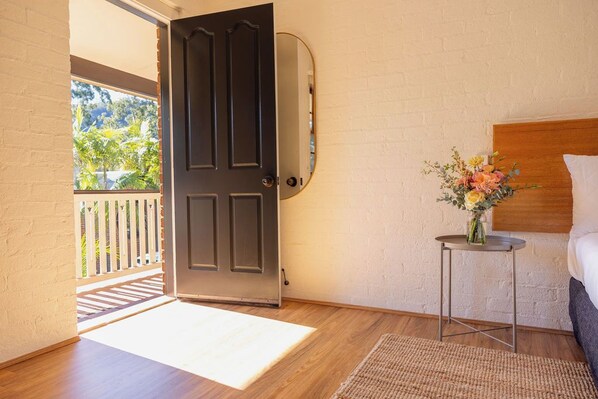 A room with a wooden floor, a dark door, a small round table with a vase of flowers, and a bed with a wooden headboard. - The Retreat at Wisemans (Sydney)