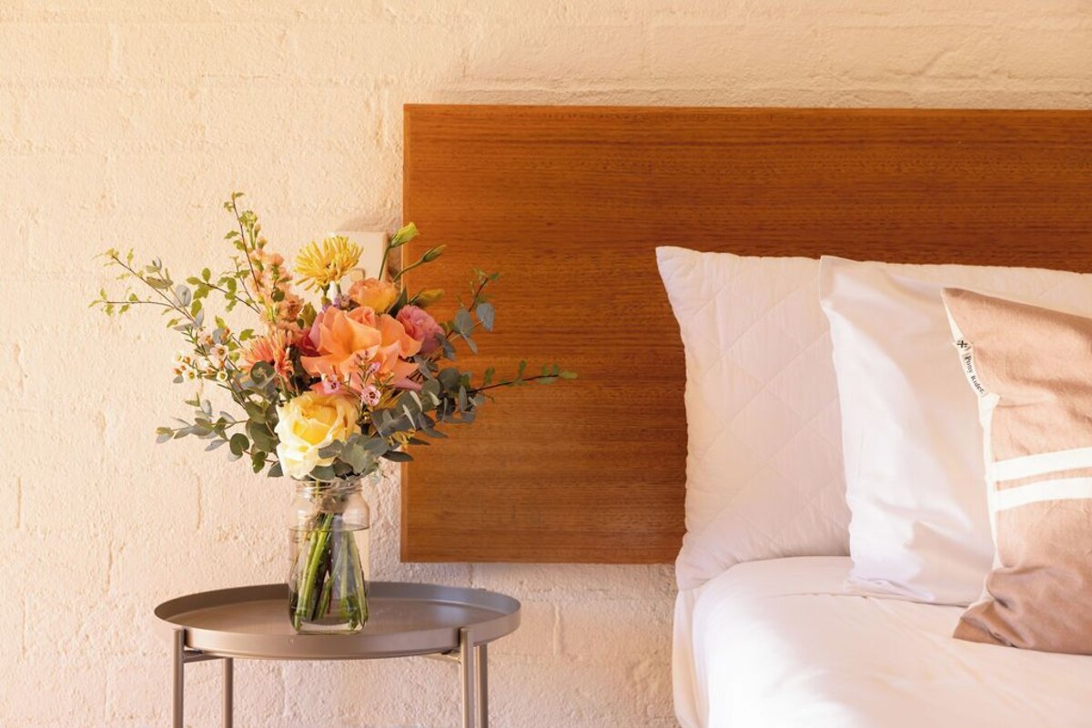 Een bed met wit beddengoed en een houten hoofdbord, een klein rond tafeltje met een vaas bloemen, en een effen muur op de achtergrond.
