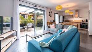 Un salon moderne avec un canapé bleu, une table basse et un téléviseur posé sur un meuble en bois. Une porte-fenêtre coulissante donne accès à un patio aménagé avec du mobilier d’extérieur. On aperçoit la cuisine à l’arrière-plan, équipée d’appareils électroménagers modernes.