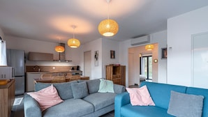 Un salon moderne avec un canapé gris et un canapé bleu, tous deux agrémentés de coussins. Deux suspensions sont suspendues au plafond. On aperçoit à l’arrière-plan une cuisine avec des meubles en bois et un coin repas composé d’une table et de chaises.