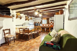 A rustic kitchen with wooden beams, a dining table with chairs, a green sofa, and a tiled floor.