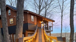 Un pont en bois, illuminé de guirlandes lumineuses, mène à un chalet en bois doté d’un balcon donnant sur une étendue d’eau.