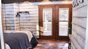 Une chambre confortable avec un plancher en bois, un lit recouvert d’une couvre-lit bleu foncé, une porte en bois et un petit tapis blanc.