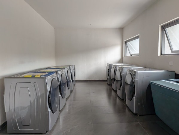 A laundry room with multiple washing machines and dryers, a tiled floor, and windows allowing natural light.