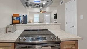 Een moderne keuken met een roestvrijstalen fornuis, een inbouwoven en een magnetron. Er is een plafondventilator, een raam met zonwering en een deur met een handgreep.