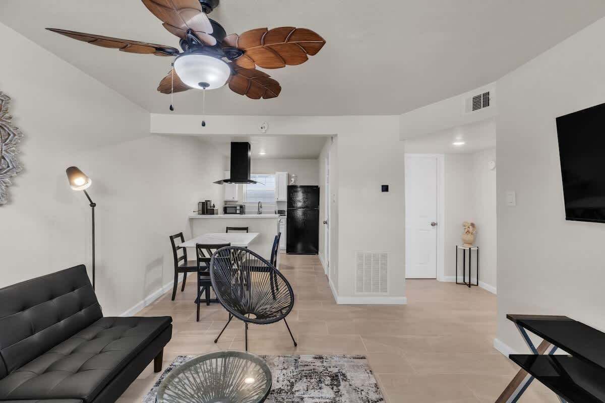 Un soggiorno moderno con un divano nero, un'area pranzo con tavolo e sedie, un ventilatore a soffitto e una cucina sullo sfondo.