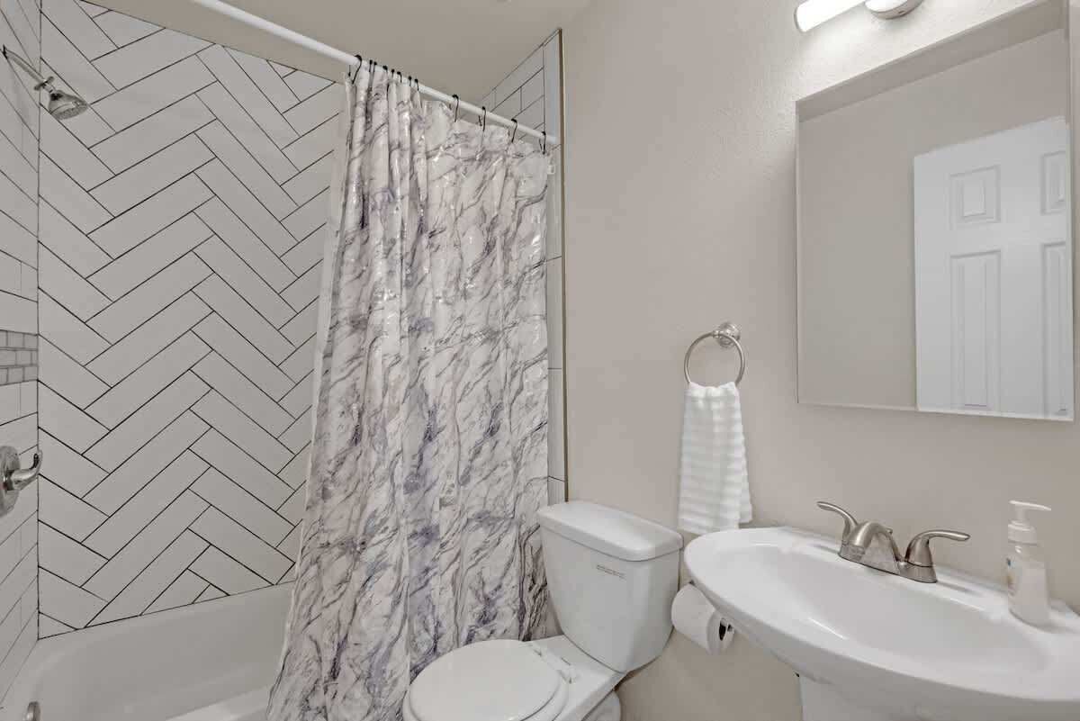 Une salle de bain avec un carrelage disposé en motif «chevrons», un rideau de douche, des toilettes, un lavabo avec un miroir et un porte-serviettes.
