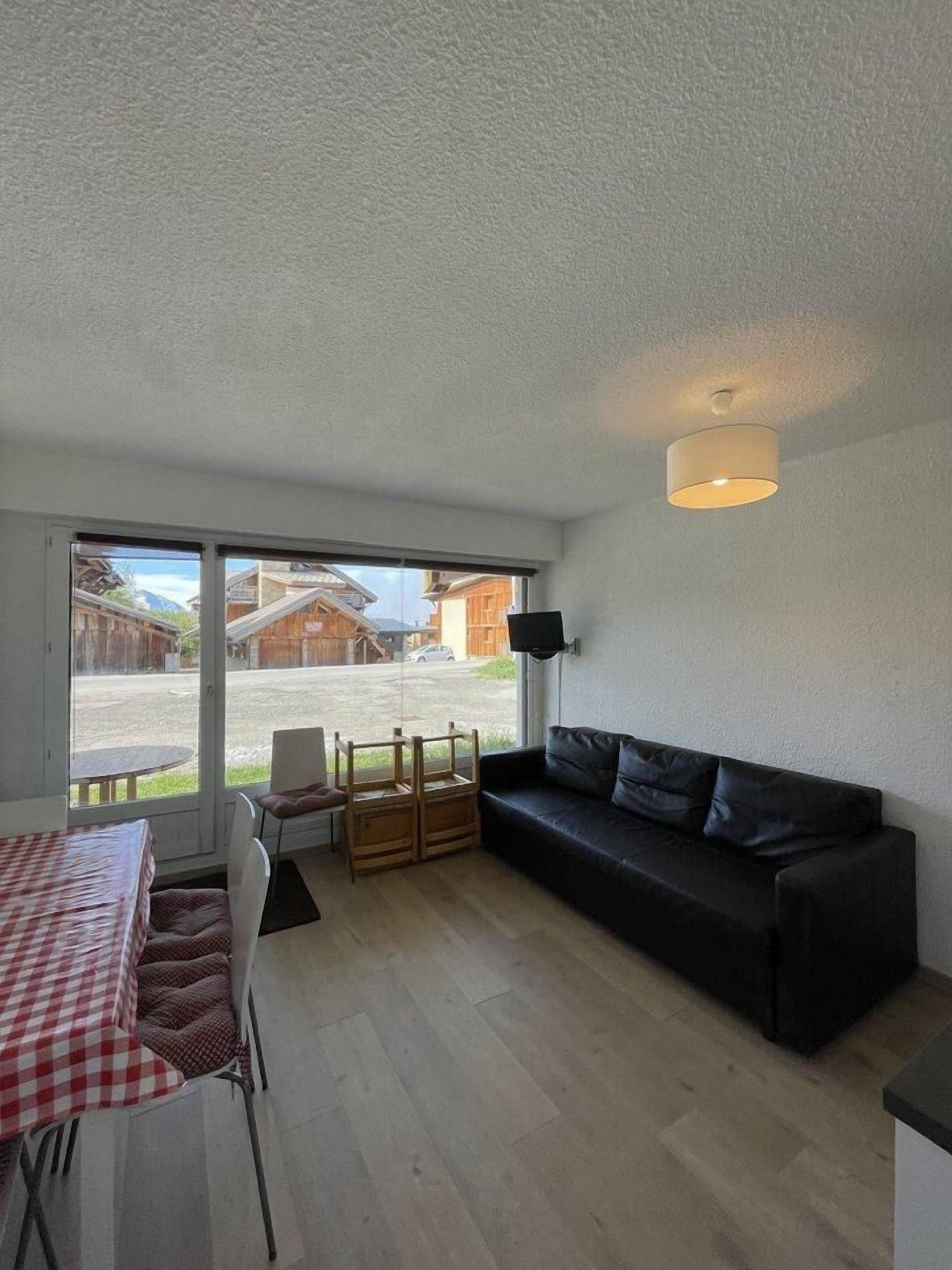 Una habitación con un sofá negro, una mesa de comedor con mantel a cuadros rojo y blanco, y vistas a una calle con edificios y un coche.