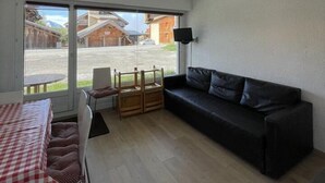 A room with a black sofa, a dining table with a red and white checkered tablecloth, and a view of a street with buildings and a car. - 350m from ski slopes, Ground floor, terrace, tv, 24m², Alpe d'Huez (L'Alpe d'Huez)