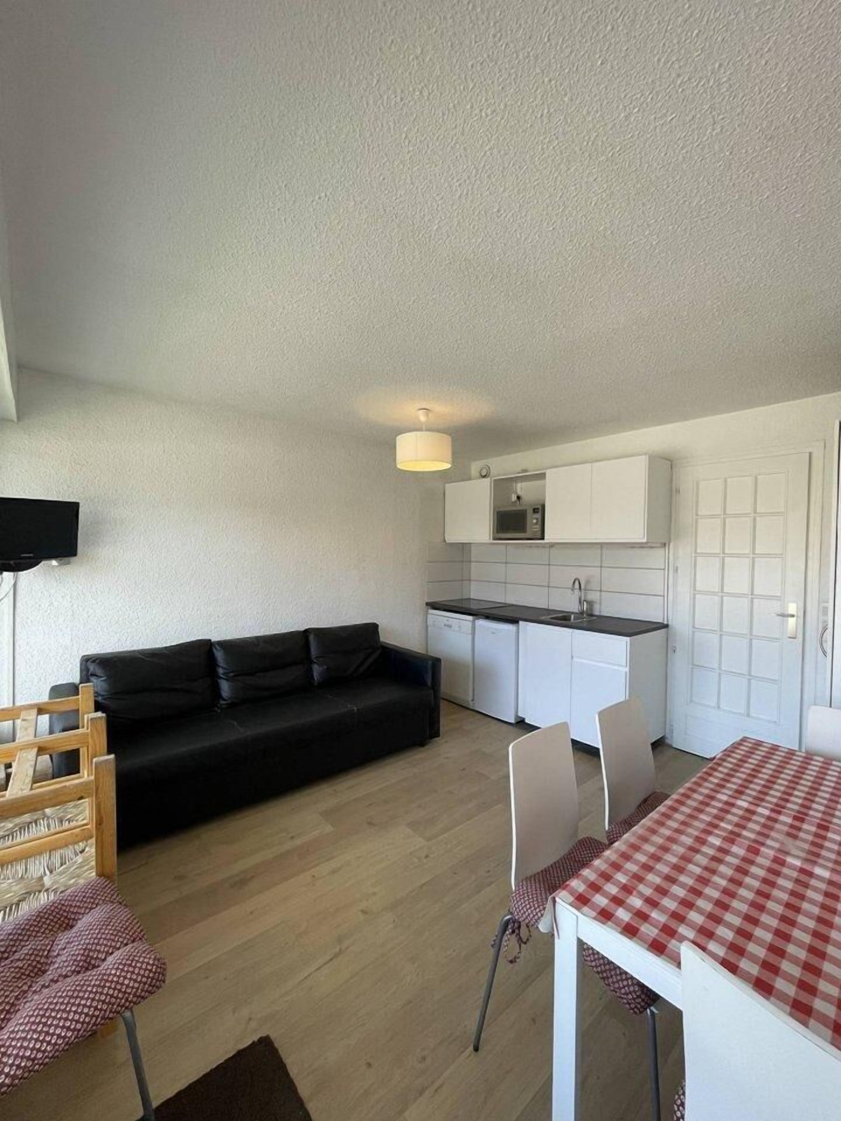 Ein kompakter Wohnraum mit Küchenzeile, einem schwarzen Sofa, einem Esstisch mit kariertem Tischläufer und einem Holzsessel.