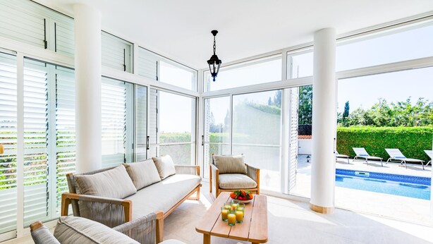 Una habitación amplia con un gran ventanal que da a una zona de piscina, un sofá, una silla y una mesa con vasos de jugo de naranja.