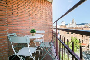 A balcony with a brick wall, a small table with a potted plant, and metal chairs. A view of a cityscape with historical buildings is visible through the railing.