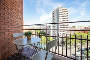 En balkon med et rundt bord, to stole, en potteplante og udsigt til en høj bygning og bybillede.