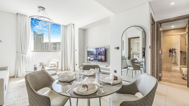 A modern hotel room with a dining area, a large window with a city view, a flat-screen TV, and a walk-in shower. - New 2BR with Pool & Gym in Green Sobha Community (Dubai)