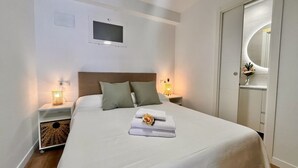 Cama bem arrumada com edredom branco e travesseiros, mesa de cabeceira com abajur e televisão na parede.