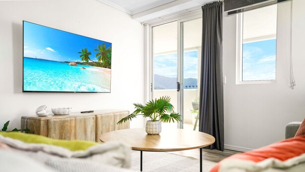 A living room with a large flat-screen TV on the wall, a round wooden coffee table with a potted plant, a sofa, and a sliding glass door leading to a balcony.