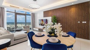 A modern living room with a round dining table, blue chairs, and a large window offering a city view. - Ultra Luxurious! Stunning Views, 64th Floor Pool (Business Bay)