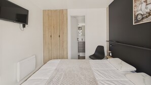 A modern bedroom with a bed, a black chair, a TV mounted on the wall, and a wooden panel wall. - Chalet for 6 - Heated Pool - Terrace (Bretignolles-sur-Mer)