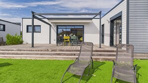 A modern house with a patio area featuring two lounge chairs, a wooden deck, and a glass door leading to an interior dining area. - Chalet for 6 - Heated Pool - Terrace (Bretignolles-sur-Mer)