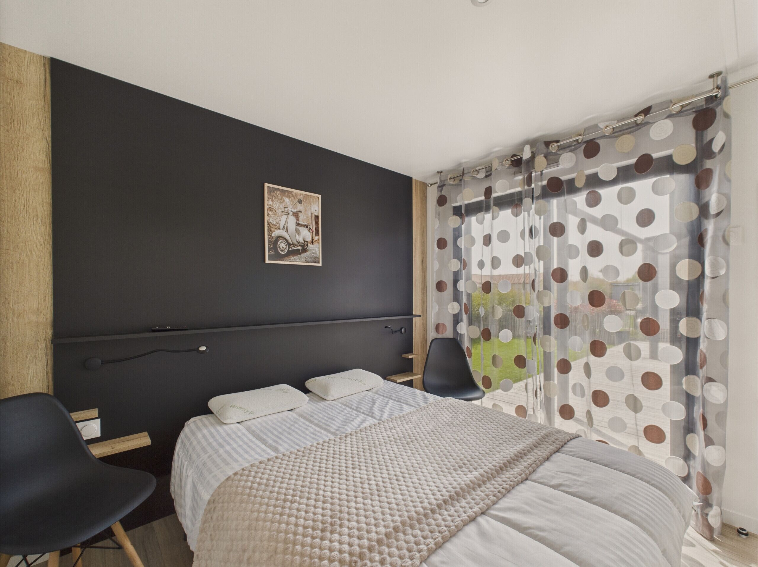 A bedroom with a bed, two chairs, a wooden headboard, a polka dot curtain, and a framed picture on the wall.