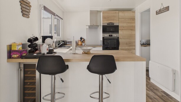 A modern kitchen with white cabinetry, a wooden countertop, two black bar stools, and a built-in oven. - Chalet for 6 - Heated Pool - Terrace (Bretignolles-sur-Mer)