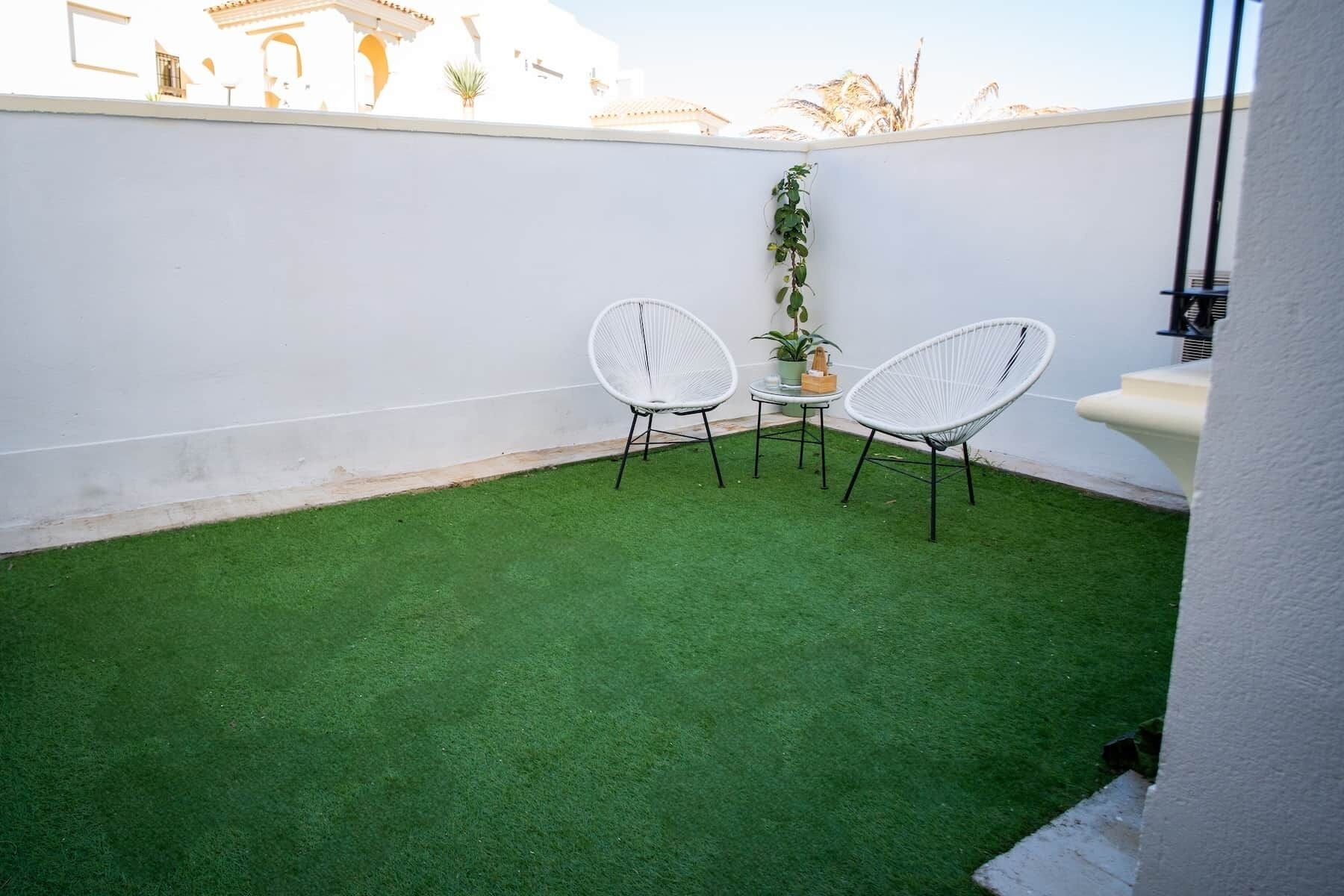 Une petite terrasse avec un gazon synthétique vert, deux chaises en osier blanches et une petite table sur laquelle est posée une plante.