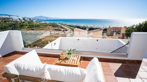 Un balcon avec vue sur la mer, une table en bois agrémentée d’une plante et une chaise blanche avec un coussin.