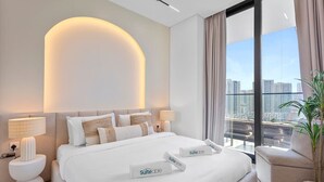 A modern hotel room with a large bed, two bedside tables with lamps, a city view, and a potted plant. - Exclusive 1 Bedroom  in Trillionaire Residences (Dubai)