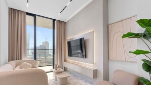 A modern living room with a large TV, comfortable seating, a view of the city, and a balcony with a plant. - Exclusive 1 Bedroom  in Trillionaire Residences (Dubai)
