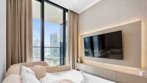 A modern hotel room with a large bed, a flat-screen TV mounted on the wall, and a city view through large windows. - Exclusive 1 Bedroom  in Trillionaire Residences (Dubai)
