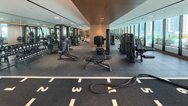 A modern gym with various exercise equipment, large windows, and a high ceiling. - Exclusive 1 Bedroom  in Trillionaire Residences (Dubai)