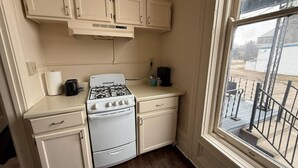 A compact kitchen with white cabinets, a gas stove, and a window overlooking a balcony. - Shearer Historical House Unit 1 (Bay City)