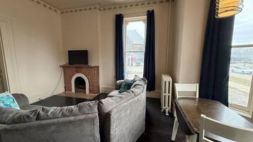 Ein Wohnzimmer mit Kamin, einer Couch, einem Esstisch mit Stühlen und Blick auf ein Gebäude durch ein Fenster.