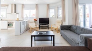 Un salon moderne avec un canapé gris, une table basse et un téléviseur. Il y a un coin cuisine avec des meubles blancs et une cuisinière. La pièce est dotée de grandes fenêtres avec des rideaux.