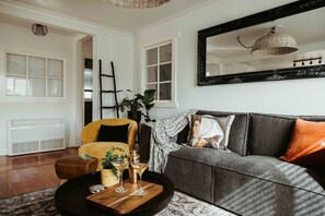 Ein Wohnzimmer mit einer Couch, einem Sessel und einem Couchtisch. An der Wand hängt ein großer Spiegel und in der Ecke steht eine Pflanze.
