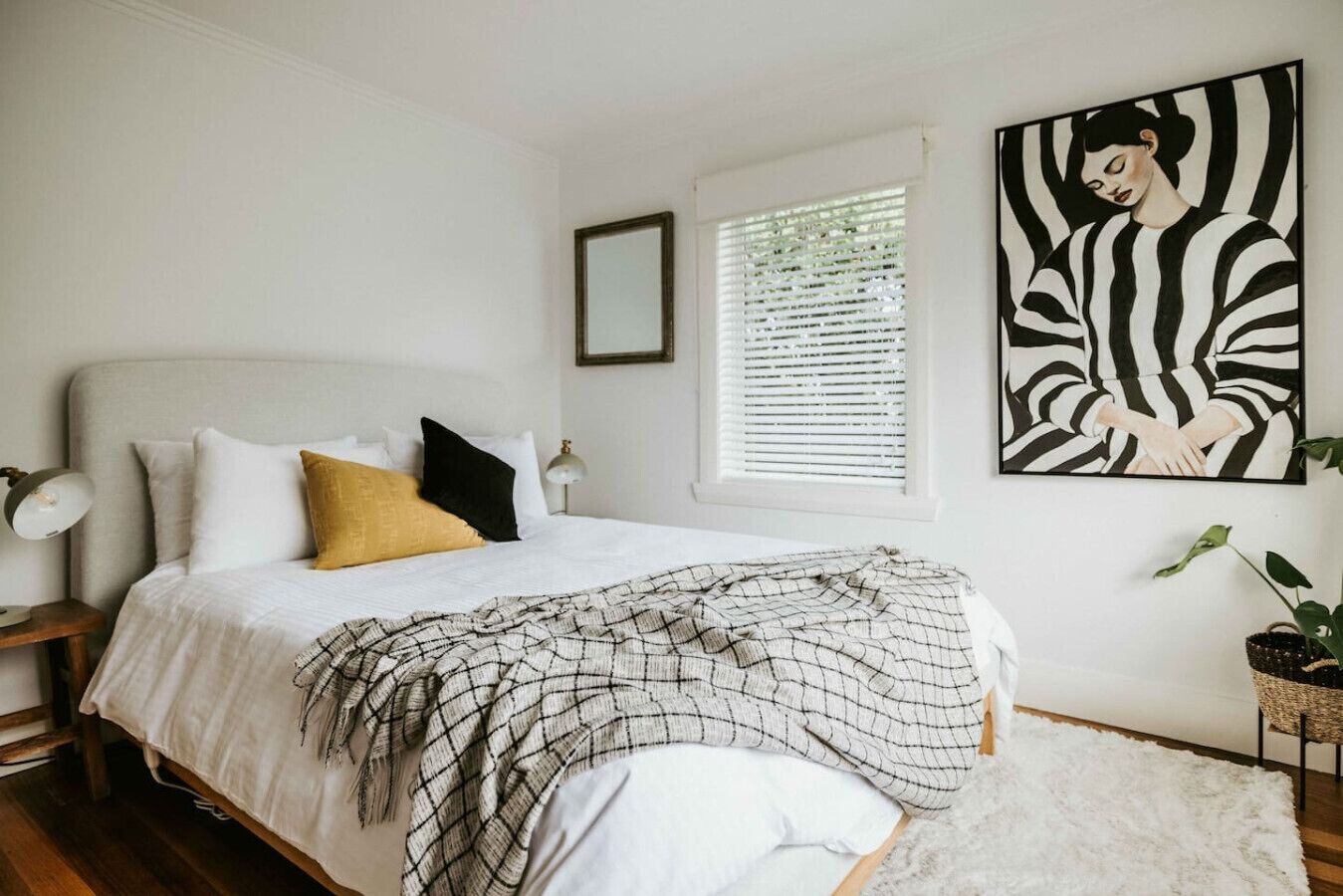 A bedroom with a bed, pillows, a blanket, a window with blinds, a wall-mounted picture, and a small plant.