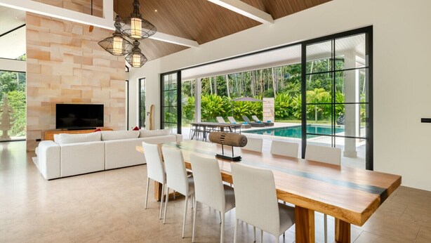 Ein modernes Wohnzimmer mit großem Fernseher, weißem Sofa, Esstisch und Stühlen. Es verfügt über eine Steinwand, große Fenster und Blick auf einen Pool.