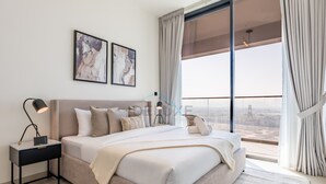 A modern bedroom with a large bed, a nightstand, a desk, and a view of the city through large windows. - Skyline-View 1BR with Balcony,Minutes to Downtown and Marina, Binghatti Onyx, JVC (Dubai)