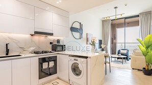 A modern kitchen with a built-in washer and dryer, a microwave, and a large window with curtains. - Skyline-View 1BR with Balcony,Minutes to Downtown and Marina, Binghatti Onyx, JVC (Dubai)