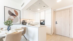 A modern kitchen with white cabinetry, a central island, and a dining area with a table set for four. - New 1 BR with 2 Balconies Near Circle Mall Luma22 West Tower (Dubai)
