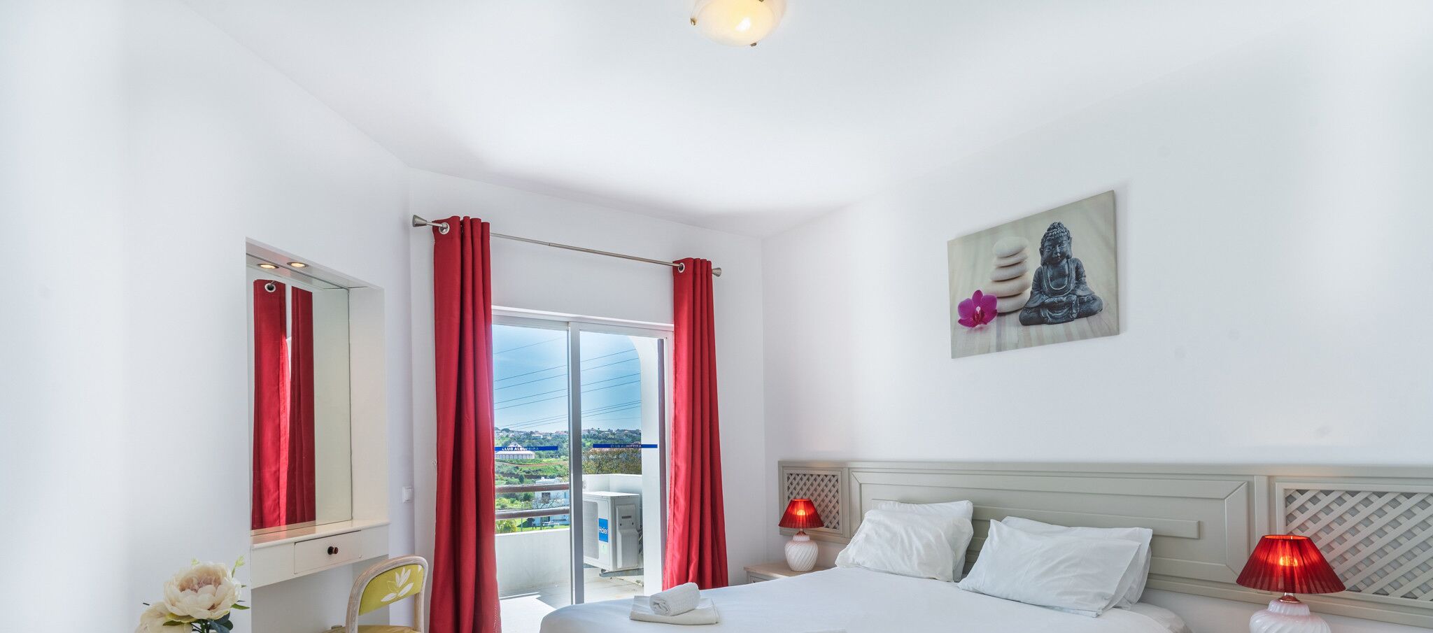 A hotel room with a bed, a nightstand, a lamp, a window with red curtains, a mirror, and a Buddha statue on the wall.