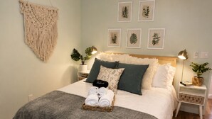 Una cama bien hecha con un edredón gris, un colgador de macramé y láminas botánicas enmarcadas en la pared.