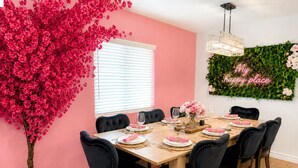 Comedor con mesa de madera para seis personas, sillas tapizadas en negro, un árbol artificial rosa grande y un adorno de follaje verde montado en la pared con un letrero de neón.