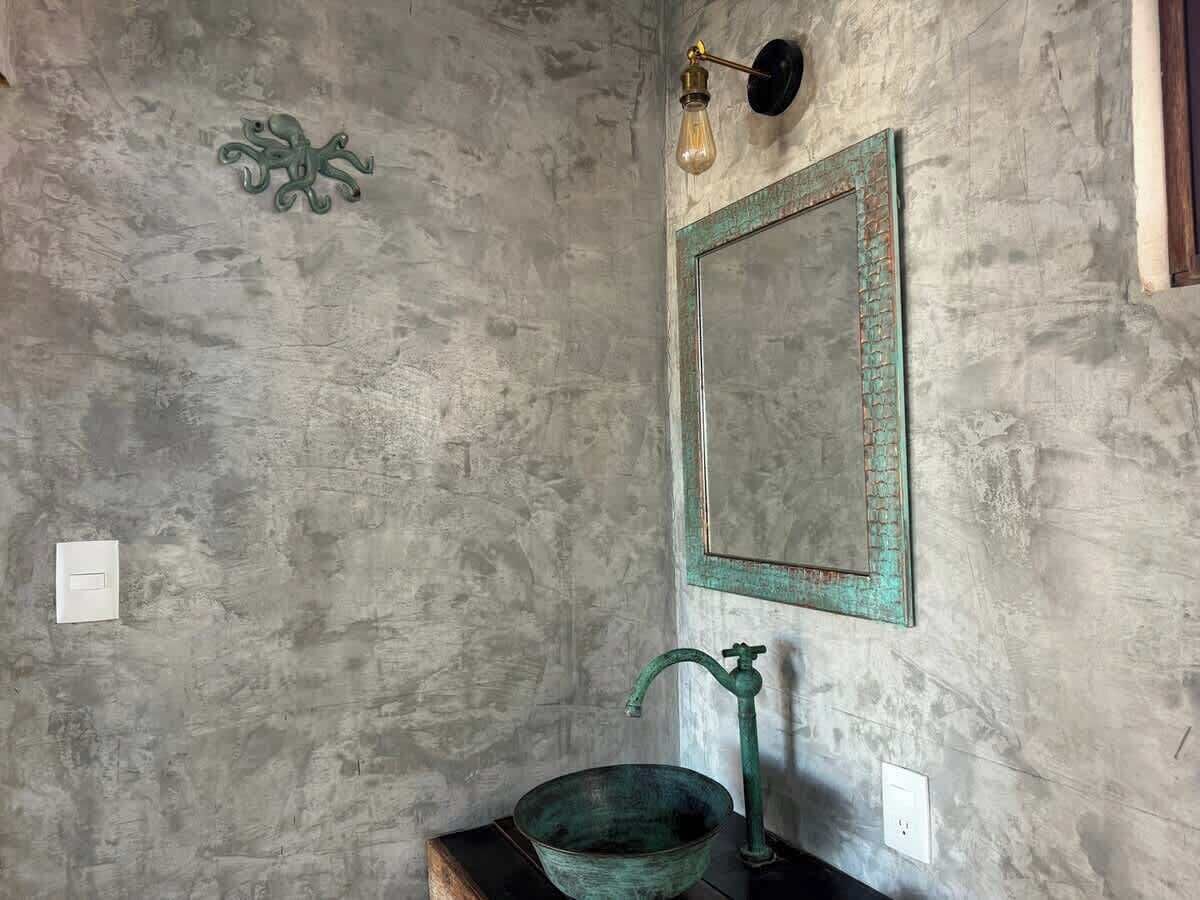 A bathroom with a rustic concrete wall, a green metal sink, a mirror with a green frame, and a light fixture.
