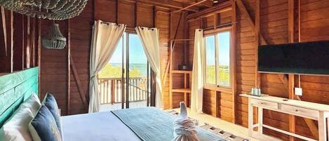 Een slaapkamer in een houten chalet, met een bed, gordijnen, een plafondventilator, een televisie en een balkon met uitzicht.