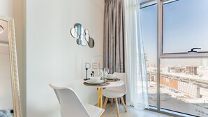Una habitación moderna con una mesa de comedor, sillas blancas, un ventanal con vistas a un paisaje urbano, un espejo en la pared y un sofá azul capitonado.