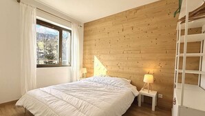 Un dormitori amb paret de fusta, un llit amb roba de llit blanca, una tauleta de nit amb una làmpada i una finestra amb cortines.