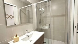 Bany modern amb dutxa tancada amb vidre, un lavabo blanc amb calaixera de fusta i un mirall gran.